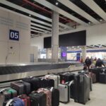 Baggage Claim Area 5 at Jorge Chávez Airport with a large amount of luggage on the ground, an empty conveyor belt, and a few passengers visible in the background.