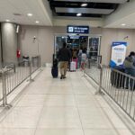 View from inside Jorge Chávez Airport looking toward the exit from Customs into the arrivals hall, with passengers walking through. Bilingual signage reading "Aduanas" and "Customs" is visible on the right side.