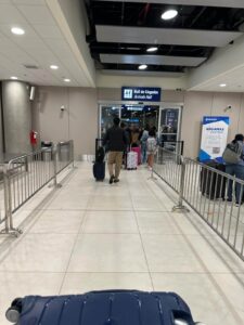 View from inside Jorge Chávez Airport looking toward the exit from Customs into the arrivals hall, with passengers walking through. Bilingual signage reading "Aduanas" and "Customs" is visible on the right side.
