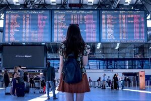 Woman seen from behind, appearing slightly unsure, looking at arrivals and departures boards inside an airport terminal.