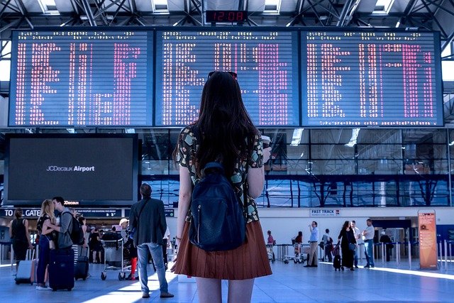 Woman seen from behind, appearing slightly unsure, looking at arrivals and departures boards inside an airport terminal.